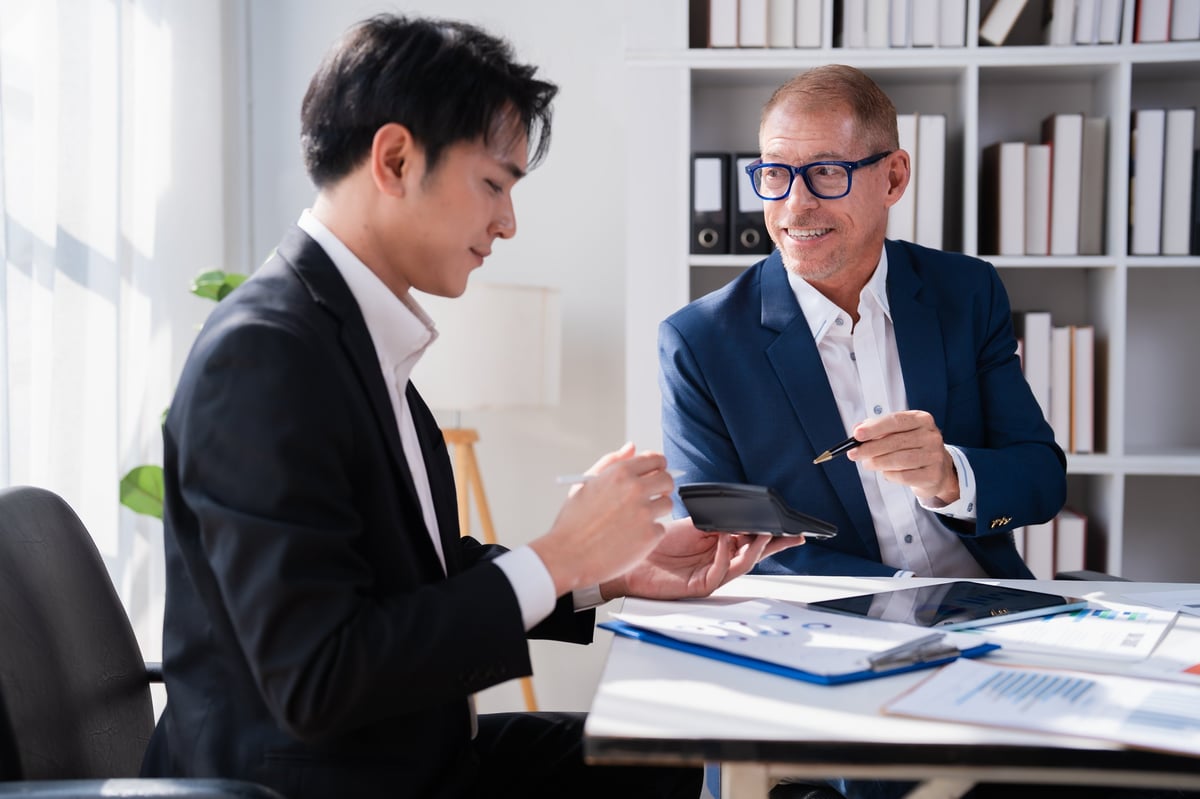 Zwei Geschäftsmänner arbeiten gemeinsam an einem Finanzplan, prüfen Dokumente und nutzen einen Taschenrechner, während sie Investitionsstrategien in einem hellen, zeitgenössischen Büro besprechen.